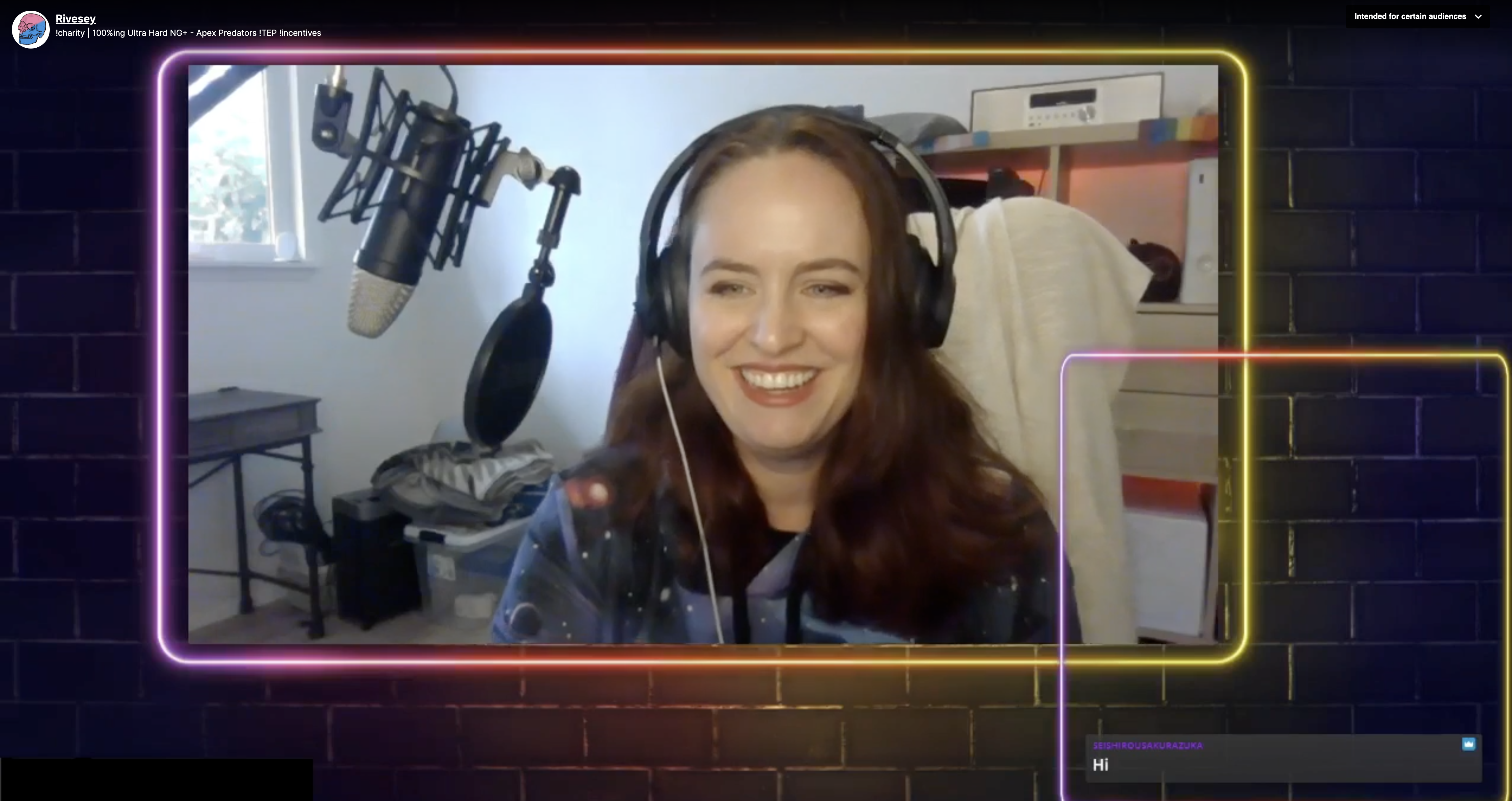 Twitch Streamer Rivesey is a light skinned person with blue eyes and long reddish brown hair, and is seen smiling while wearing black headphones and sitting infront of a microphone